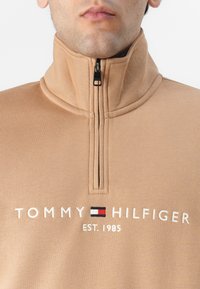 Béžová mikina s krátkym zipsom z jemného materiálu s vysokým golierom. Obsahuje vyšívaný nápis "TOMMY HILFIGER" a rok založenia, doplnený logom.