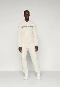 Bež trenirka s kapuco in modrim logotipnim trakcem, značilna po ohlapnem kroju in rebrastih manšetah, v kombinaciji z belimi supergami.