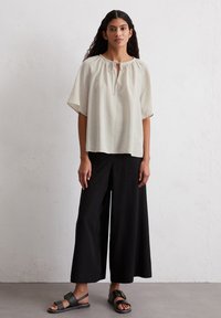 Elfenbeinfarbenes kurzärmliges Blusenshirt mit gerafftem Ausschnitt, kombiniert mit weiten schwarzen Hosen und schwarzen Sandalen mit einer Schnallenapplikation.