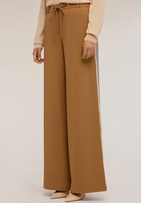 Brede bruine broeken met zijstreep in wit en zwart, elastische tailleband met trekkoord, gedragen met een beige langemouwen top en puntige naaktkleurige hakken.