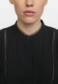 Schwarze Bluse mit vertikalen Falten, rundem Kragen mit dekorativer Spitzenbordüre und einem einzelnen Knopfverschluss am Hals.