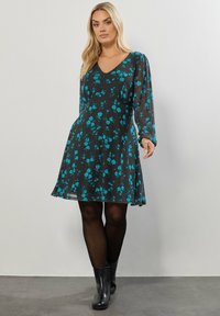 Schwarzes Kleid mit langen Ärmeln und V-Ausschnitt, mit einem türkisfarbenen Blumenmuster. A-Linie, semitransparenter Stoff, kombiniert mit schwarzen Knöchelstiefeln.