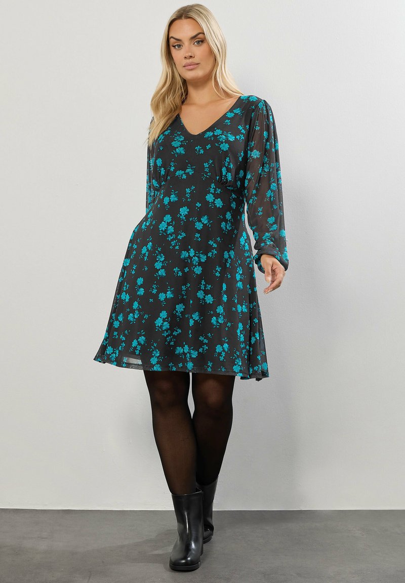 Schwarzes Kleid mit langen Ärmeln und V-Ausschnitt, mit einem türkisfarbenen Blumenmuster. A-Linie, semitransparenter Stoff, kombiniert mit schwarzen Knöchelstiefeln.
