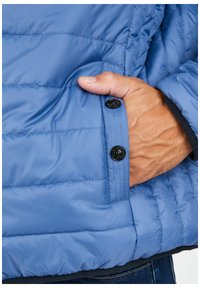 Hellblaue Steppjacke mit strukturiertem Material, in die eine Hand in eine Seiten- tasche ruht, die mit zwei schwarzen Knöpfen gesichert ist.