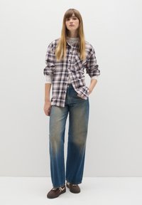 Camicia a quadri bianca, nera e rosa, indossata sopra una maglia in pizzo. I jeans blu sbiaditi a gamba larga e le sneakers marroni completano il look.