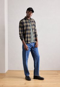 Cămașă în carouri verzi și negre, tricou verde solid dedesubt, blugi din denim albastru, pantofi negri și o șapcă neagră, stând pe un podea din lemn.