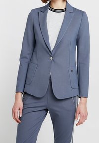 Dopasowana marynarka z miękkiego, niebieskiego materiału. Do jej szczególnych cech należą dwie przednie kieszenie, zapięcie na jeden guzik oraz klapa w stylu notch.