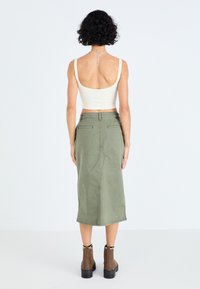Olive green, mid-length skirt made of cotton with a smooth texture, featuring back pockets and a straight fit. Worn with a fitted, light top.
