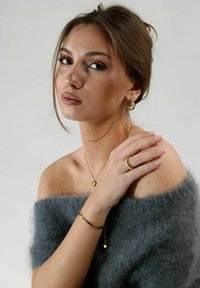 La joyería de oro incluye pendientes de aro, un collar con colgante en forma de corazón y un anillo texturizado. La modelo lleva un suéter suave, gris y sin hombros.