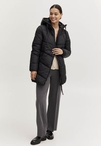 Svart puffjacka med huva, quiltad design, dragkedja framtill, bärs över en beige tröja och grå vida byxor. Skorna är svarta loafers.