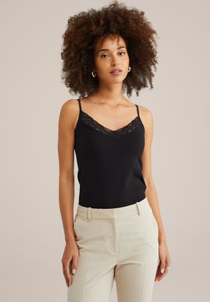 Donna con capelli ricci che indossa un top in pizzo nero e pantaloni beige, in piedi contro uno sfondo chiaro e semplice.