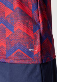 Rød og blå mønstret treningstrøye med en teksturert, sikksakkdesign. Inkluderer "DRYCELL"-logo nær kanten.