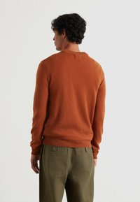 Sweter w kolorze rdzy z teksturowanym wykończeniem, okrągłym dekoltem i ściągaczami, w połączeniu z oliwkowymi spodniami.