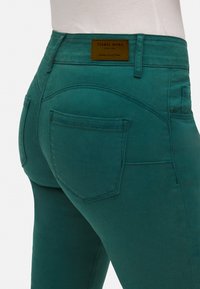 Jeans de mezclilla turquesa con cintura alta, que cuentan con dos bolsillos traseros, costura sutil y una etiqueta de cuero marrón con el nombre de la marca.