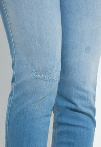 Jeans in denim blu chiaro con sottili patch consumati nella zona delle cosce, primo piano che mostra la texture del tessuto e i dettagli delle cuciture.