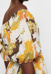 Kwiecista sukienka odkrywająca ramiona, wykonana z lekkiego, teksturowanego materiału. Żywy wzór kwiatowy w kolorach żółtym, pomarańczowym i zielonym z bufkami na rękawach.