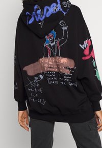 Persona con sudadera negra que tiene dibujos y texto en estilo graffiti colorido, incluyendo una figura con cabello azul y frases como "por favor, intenta de nuevo."