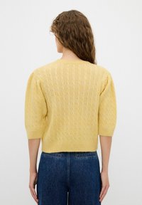 Maglione giallo lavorato a maglia con un motivo a trecce testurizzato, maniche corte a palloncino e scollatura rotonda. Indossato con jeans blu a vita alta.