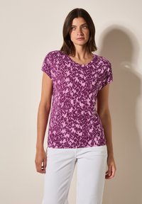 Top de manga corta en un patrón abstracto de púrpura y rosa, con escote redondo, fabricado en un tejido suave y de corte relajado, combinado con pantalones blancos.