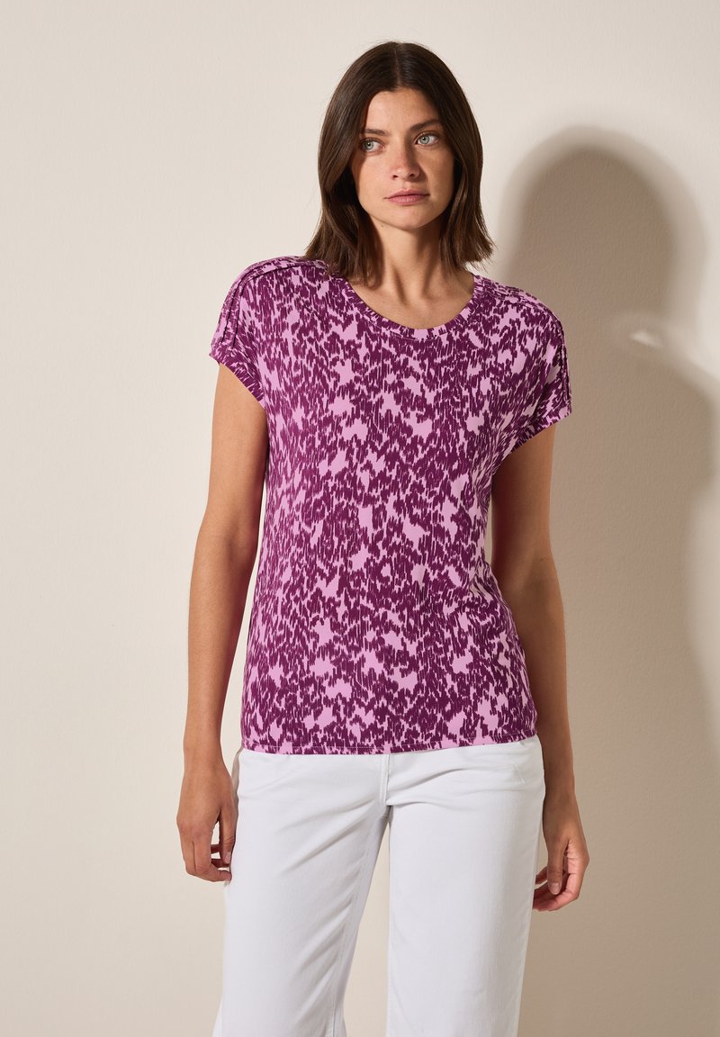 Top de manga corta en un patrón abstracto de púrpura y rosa, con escote redondo, fabricado en un tejido suave y de corte relajado, combinado con pantalones blancos.