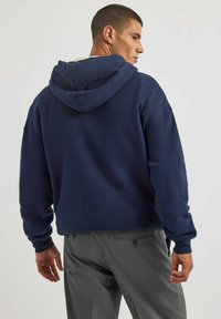 Granatowa bluza z kapturem o luźnym kroju, grubej teksturze materiału i ściągaczach. Noszona z szarymi, dopasowanymi spodniami, podkreślającymi swobodny styl.