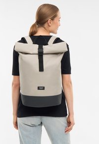 Sac à dos roll-top en beige et noir, fabriqué en tissu imperméable. Présente des bretelles réglables et une boucle avant pour une fermeture sécurisée.