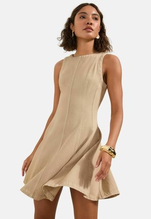 Frau mit lockigem Haar, die ein ärmelloses beiges Kleid und goldenen Schmuck trägt, posiert vor hellem, einfarbigem Hintergrund.