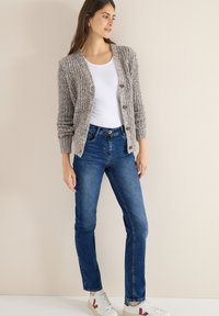 Pletený šedý kardigan s knoflíkovou přední částí, bílé tričko, modré džíny a bílé tenisky s barevnými detaily. Hladká textura a volný střih.