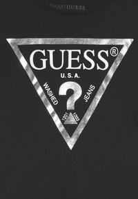 Must musta mustaga, millel on suur hõbedane kolmnurkne graafika. Keskosas on "GUESS" logo, "?" ja "WASHED JEANS" tekst julgete valgete tähtedega.