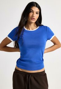 Blå kortærmet t-shirt med en hvid ribstrikket rund halsudskæring og ærmekanter. Glat stof med en tætsiddende pasform. Kombineret med mørkebrune bukser.