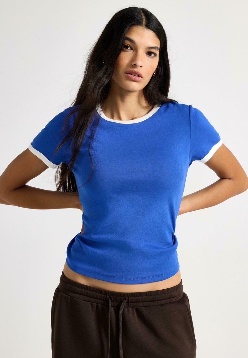 Blå kortærmet t-shirt med en hvid ribstrikket rund halsudskæring og ærmekanter. Glat stof med en tætsiddende pasform. Kombineret med mørkebrune bukser.