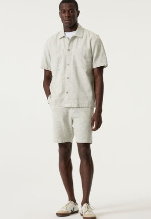 Lichtgroen linnen overhemd met korte mouwen en twee borstzakken, gecombineerd met bijpassende korte broek. Model draagt casual sneakers.