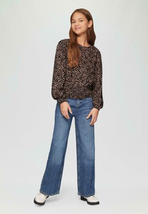 Schwarzes, blumiges Langarmoberteil mit elastischem Saum, kombiniert mit blauen, weiten Jeans. Das Model trägt cremefarbene Schuhe mit schwarzen Sohlen.