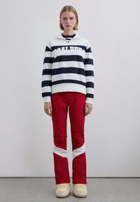 Jersey de rayas blancas y azul marino con cuello con cremallera de tres cuartos y texto destacado, combinado con pantalones rojos con detalles diagonales blancos y botas amarillas.