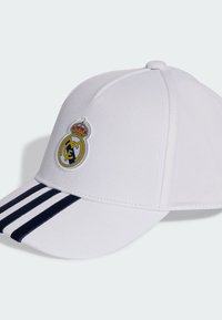 White cap featuring a rounded crown, a navy embroidered crest with a crown, and three navy stripes on the visor. Smooth fabric texture.