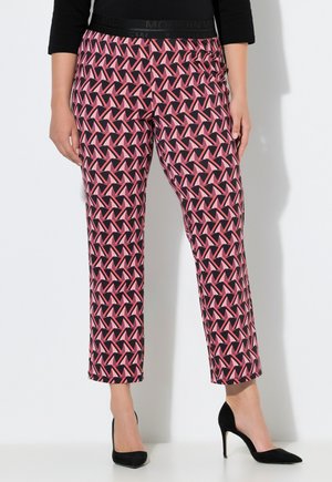 Frau trägt gerade geschnittene Hosen mit pink-schwarzem geometrischem Muster und schwarze High Heels, steht vor weißem Hintergrund.