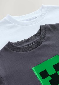 Deux t-shirts à col ras du cou : l'un blanc et l'autre gris foncé. Le t-shirt gris présente un motif vert en pixels. Cols côtelés et texture douce.