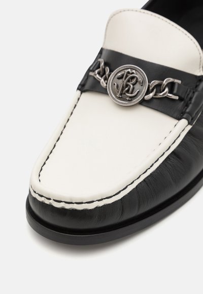Black and white leather loafer with a rounded toe, featuring a silver chain accent and logo medallion on a black strap. Visible stitching detail.