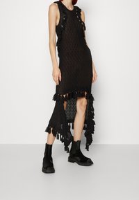 Svart stickad klänning med ärmlös design, fransdetaljer, hög-låg nederkant och öppen virkning. Kombinerad med svarta ankelboots.