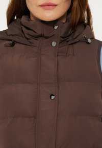 Femme portant une doudoune marron sans manches à col montant et boutons-pression, visible du menton à la taille sur un fond uni.