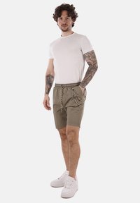 Pantaloni corti in tessuto verde oliva con elastico in vita, cordoncino decorativo, tasche laterali, abbinati a una t-shirt bianca e sneakers bianche.