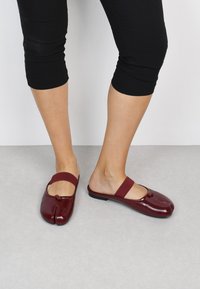 Zapatos de charol burdeos sin cordones, con puntera redondeada, tira elástica y detalle de nudo decorativo en la parte superior.