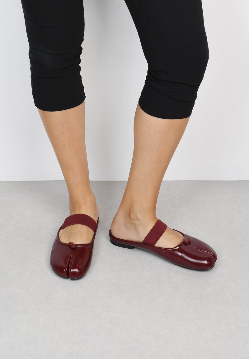 Zapatos de charol burdeos sin cordones, con puntera redondeada, tira elástica y detalle de nudo decorativo en la parte superior.