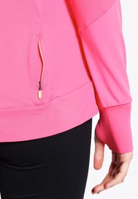 Top atlético ajustado de color rosa, hecho de una tela suave; cuenta con un bolsillo con cremallera y agujeros para los pulgares en los puños para funcionalidad y comodidad.