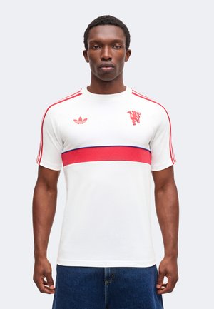 Homme debout face à l’avant portant une chemise blanche à manches courtes avec une bande horizontale rouge et un emblème de lion rouge, associée à un jean bleu.