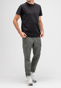 Schwarzes Baumwoll-T-Shirt mit Rundhalsausschnitt, kombiniert mit olivegrünen Cargo-Hosen. Die Hosen verfügen über Seitentaschen und sind an den Bündchen umgeschlagen. Sneaker mit gemustertem Design.
