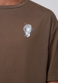 Brūna kokvilnas t-krekls ar izšūtu grafiku, kas attēlo klasisku krūšautu skulptūru ar čūskas akcentiem pelēkā, melnā un sarkanā krāsā.