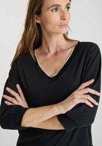 Donna con lunghe capelli castani, indossa una maglietta nera con scollatura a V e bordi dorati, braccia incrociate, guarda di lato su uno sfondo semplice.