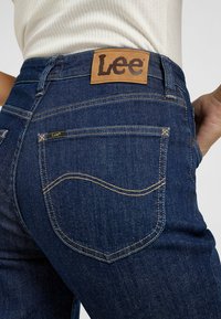 Jeans de mezclilla azul oscuro con una etiqueta de cuero que dice 'Lee' en la cinturilla, con costuras doradas y un diseño de bolsillo trasero curvado.