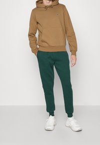 Brun luvtröja med framficka och gröna joggers. Vita sneakers kompletterar outfiten, som har en avslappnad passform och stickade muddar.